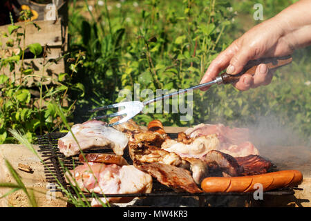 Barbecue. Saucisses, cuisses de poulet, des steaks de porc et d'un bacon sur un grill grille. Une vieille femme main est à l'aide d'une fourchette pour tourner un morceau de viande. Banque D'Images