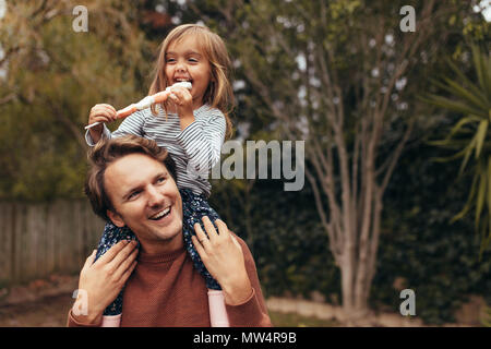 Smiling girl sitting sur les épaules de son père de manger un sucre candy stick. Père et fille de passer du temps ensemble manger du sucre candy. Banque D'Images