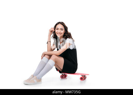 Smiling asian girl sitting on skateboard and looking at camera isolated on white Banque D'Images