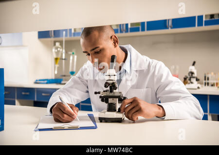 Technicien jeunes in lab coat prendre des notes tout en faisant une analyse de l'échantillon avec microscope Banque D'Images