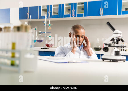 Jeune technicien pensive in lab coat assis en laboratoire et en frottant son temples Banque D'Images