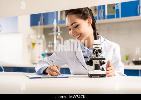 Young smiling Technicien de laboratoire prise de notes après avoir fait l'analyse de l'échantillon au microscope Banque D'Images