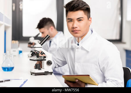 Young Scientist in lab coat assis à table en laboratoire avec microscope et digital tablet and looking at camera Banque D'Images