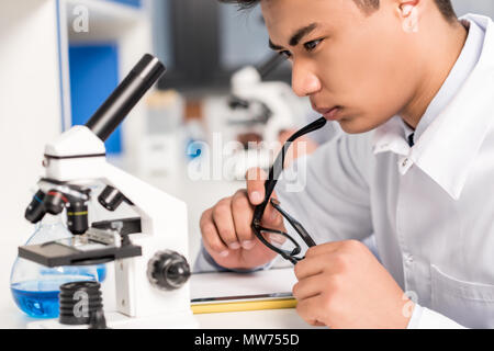 Jeune chercheur pensif assis à table en laboratoire avec microscope et tube à essai, tout en maintenant ses lunettes Banque D'Images