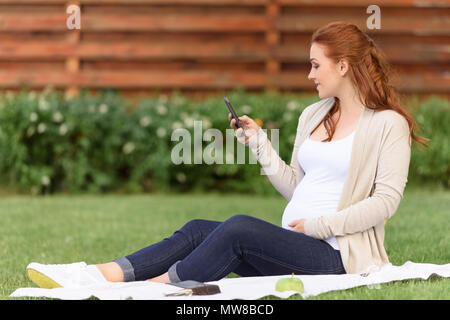 Belle rousse pregnant woman using smartphone tout en étant assis sur la pelouse verte Banque D'Images