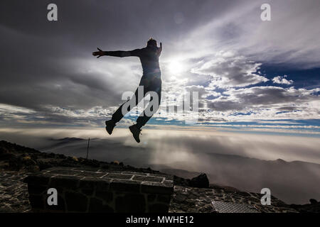 L'homme en sautant d'un banc de travail avec les mains jusqu'en face de soleil avec nuages sur ciel et sur des collines Banque D'Images