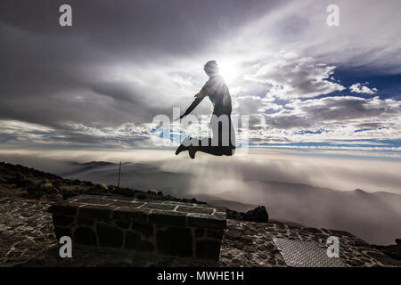 L'homme en sautant d'un banc de travail avec les mains jusqu'en face de soleil avec nuages sur ciel et sur des collines Banque D'Images