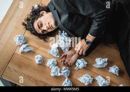 Vue de dessus de upset businessman allongé sur table avec documents froissés et looking at camera Banque D'Images