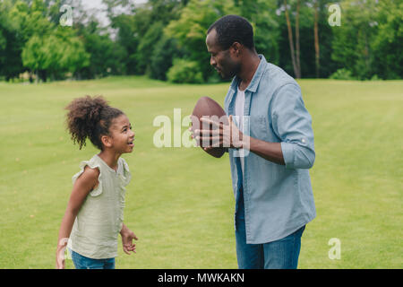 Vue latérale d'african american man et little daughter having conversation avant de jouer au football américain park Banque D'Images