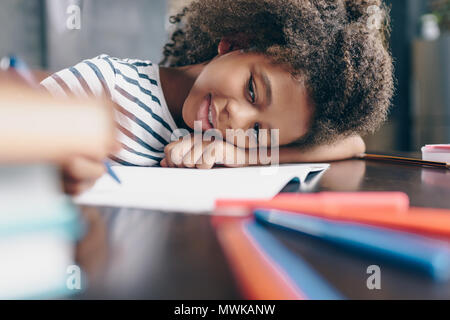 Assez petite fille assise à la table, avec sa tête sur son bras, écrit dans son cahier Banque D'Images