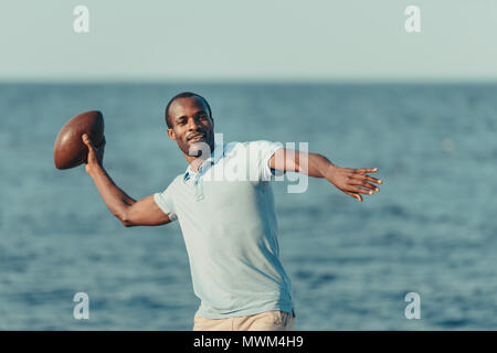 Handsome young african american man throwing rugby ball on beach Banque D'Images