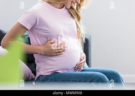 Portrait of smiling pregnant woman sitting on sofa et toucher ventre Banque D'Images