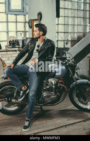 Beau jeune motocycliste en veste en cuir noire avec moto classique en atelier Banque D'Images