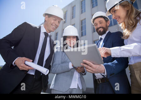 Smiling architectes professionnels dans des casques holding blueprint et using digital tablet Banque D'Images