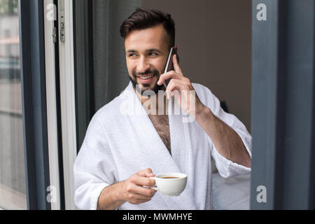 Handsome smiling barbu en peignoir talking on smartphone et de boire du café à la maison Banque D'Images