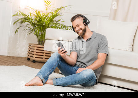 Jeune homme barbu joyeux dans les écouteurs sous smartphone et écouter la musique à la maison Banque D'Images