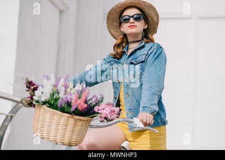 Belle fille avec chapeau, lunettes de soleil et veste en jean riding bicycle on street Banque D'Images