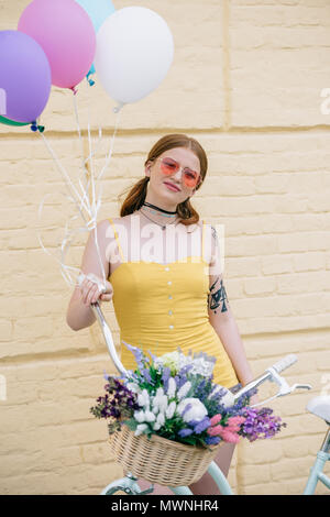 Belle jeune femme en sunglasses smiling at camera while standing avec location et de ballons colorés sur la rue Banque D'Images