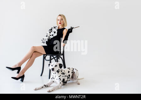 Jolie femme blonde élégante en robe noire assise sur une chaise, chien dalmatien lying on floor Banque D'Images