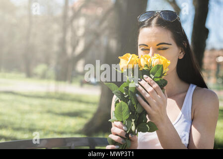 Jolie femme sentant les fleurs. Satisfait et belle fille détient des roses jaunes dans les mains, ferme les yeux, s'assoit sur un banc de parc. Cadeaux de la MOM, bi Banque D'Images