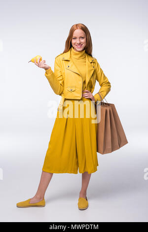 Rousse élégante fille élégante en jaune vêtements holding shopping bags et poire, isolé sur gray Banque D'Images