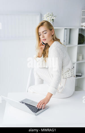 Belle concentrated businesswoman en blanc le travail avec ordinateur portable et smartphone Banque D'Images