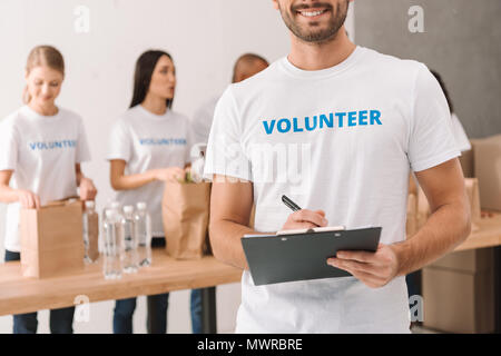 Cropped shot of volunteer écrit dans le presse-papiers avec des collègues travaillant sur l'arrière-plan flou Banque D'Images