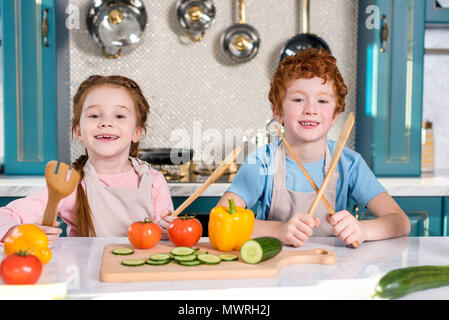 Adorables enfants holding couverts en bois et smiling at camera pendant la cuisson ensemble dans la cuisine Banque D'Images