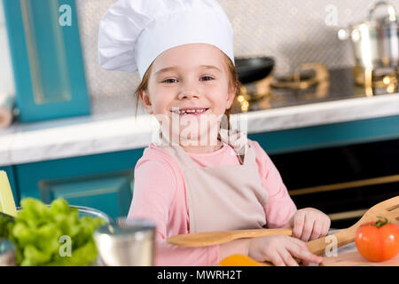 Adorable enfant en tablier et chapeau de chef smiling at camera pendant la cuisson dans la cuisine Banque D'Images