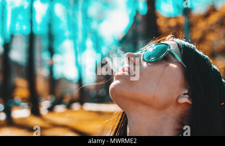 De gros plan le visage d'une femme de fumer une cigarette. Une jolie fille à lunettes est titulaire d'une cigarette dans sa bouche. Les femmes ont souligné l'arc de la nicotine satisfait Banque D'Images