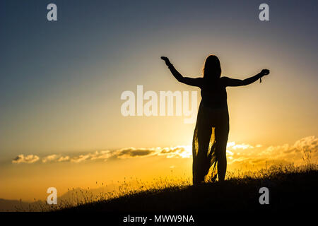 Fille en bleu transparent debout sur l'herbe verte dans les montagnes brumeuses au coucher du soleil Banque D'Images