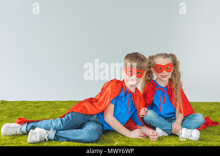 Frères et sœurs dans superhero costumes assis sur l'herbe verte et grise sur smiling at camera Banque D'Images