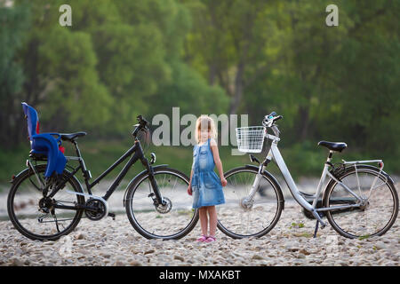 Petit très jolie jeune femme en robe bleue in front of white location avec godet et un noir avec siège enfant sur fond vert des arbres flous. Banque D'Images