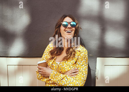 Laughing young brunette woman appuyé contre un mur dans la ville portant des lunettes de soleil et de boire une tasse de café à emporter Banque D'Images