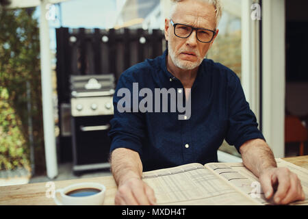 Man avec une expression grave assis à une table patio lire les pages financières dans un journal et de boire une tasse de café Banque D'Images