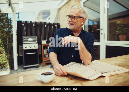 Senior alors qu'il était assis à une table à l'extérieur sur son patio lire le journal et boire une tasse de café Banque D'Images