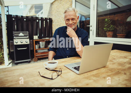 Senior man smiling satisfait alors qu'il était assis à sa table de patio de boire une tasse de café et de travailler sur un ordinateur portable Banque D'Images