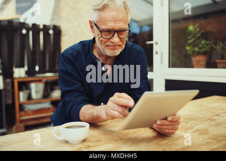 Smiling senior man drinking a cup of coffee et travailler avec une tablette numérique tout en restant assis à sa table de patio à l'extérieur Banque D'Images