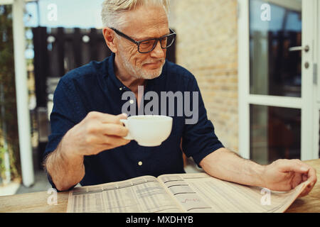 Smiling senior man sitting at table à l'extérieur sur son patio lire les pages financières dans un journal et prendre un café Banque D'Images