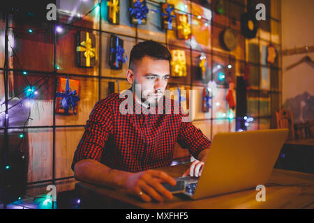 Un beau jeune homme de race blanche avec barbe et sourire à pleines dents dans une chemise à carreaux rouge fonctionne derrière un ordinateur portable gris assis à une table en bois. Les mains sur le clavier. Dans la soirée au café Banque D'Images