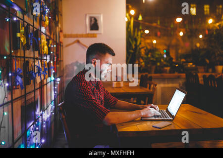 Un beau jeune homme de race blanche avec barbe et sourire à pleines dents dans une chemise à carreaux rouge fonctionne derrière un ordinateur portable gris assis à une table en bois. Les mains sur le clavier. Dans la soirée au café Banque D'Images