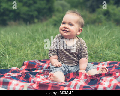 Un mignon petit bébé dans la nature est assis sur une couverture de pique-nique et rit Banque D'Images