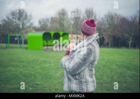 Une jeune mère est debout sur l'herbe dans un parc en hiver avec son bébé dans une écharpe de l'opérateur Banque D'Images