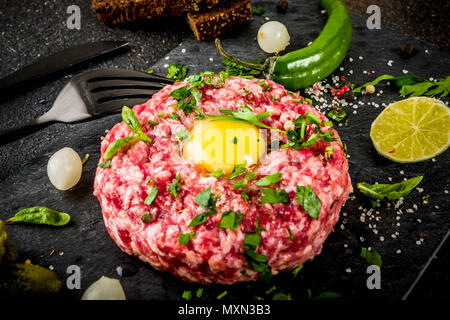 La cuisine française, avec un steak tartare de boeuf d'oeuf de caille, matières premières, cornichon et l'oignon, des herbes fraîches, du pain, l'arrière-plan foncé rusty copy space Banque D'Images