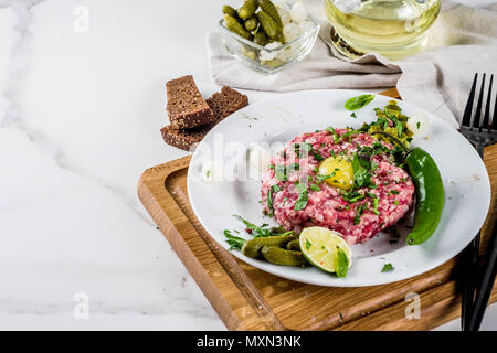 La cuisine française, avec un steak tartare de boeuf d'oeuf de caille, matières premières, cornichon et l'oignon, des herbes fraîches, du pain, du marbre blanc background copy space Banque D'Images