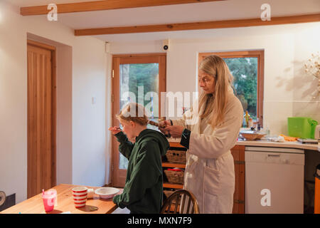 Mère de ses filles à tresser les cheveux comme elle mange son petit-déjeuner dans la cuisine. Banque D'Images