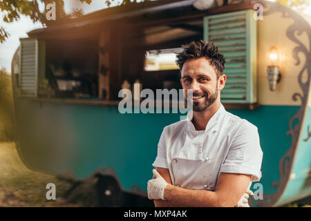 Portrait de jeune homme debout à l'extérieur cook avec son camion alimentaire en arrière-plan. Happy young camion alimentaire propriétaire. Banque D'Images