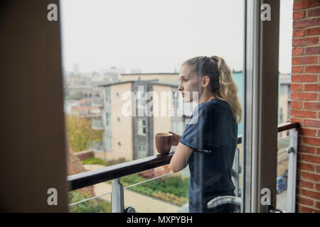 Par la fenêtre arrière, voir d'une jeune femelle adulte dans ses vêtements prendre une tasse de thé sur le balcon. Banque D'Images