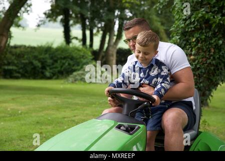 Père et fils : un père laisse son fils de 5 ans à diriger le tracteur-tondeuse qu'il coupe l'herbe Banque D'Images