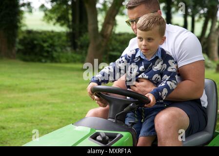 Père et fils : un père laisse son fils de 5 ans à diriger le tracteur-tondeuse qu'il coupe l'herbe Banque D'Images